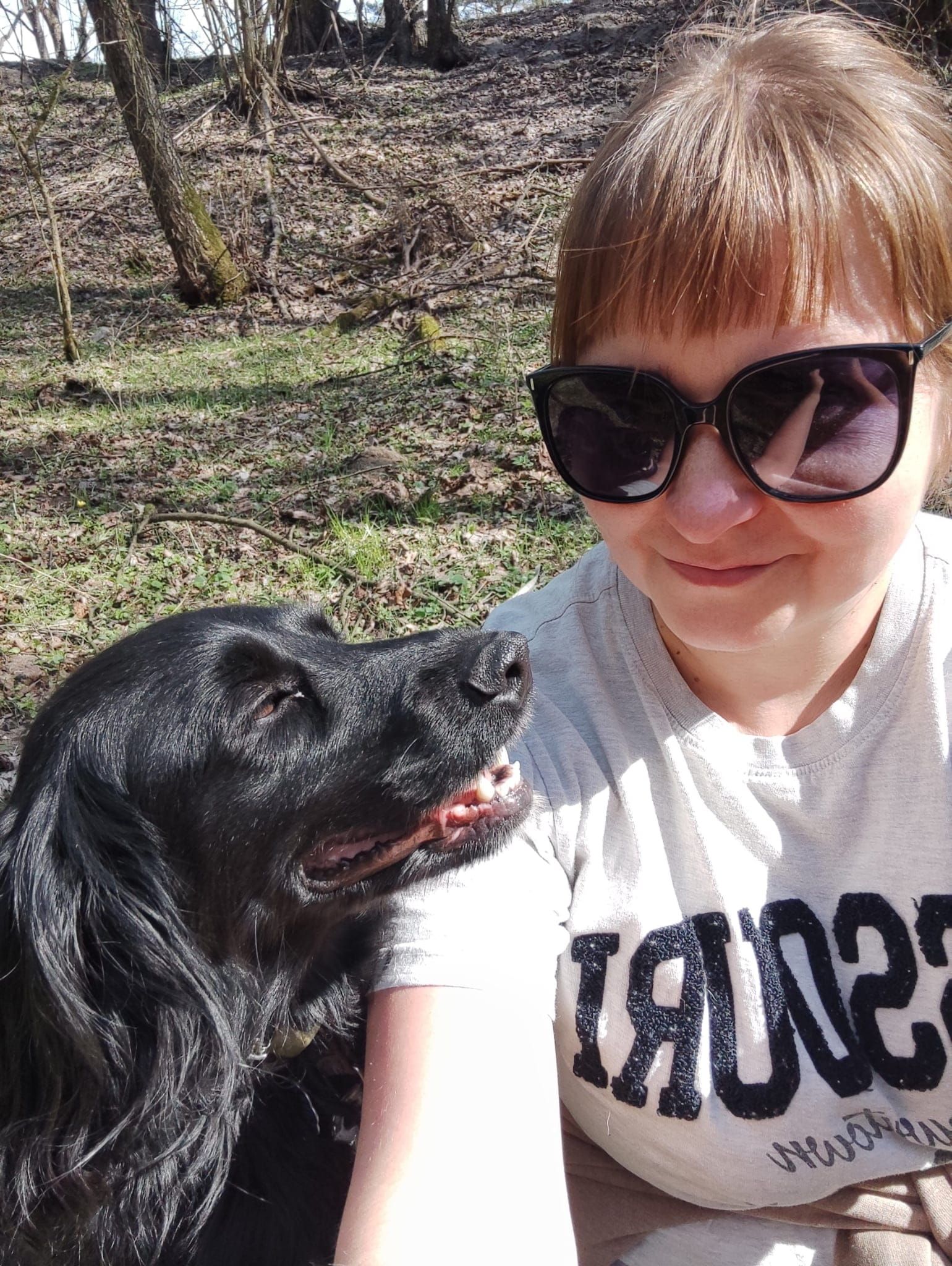 Czarny spaniel z uniesioną mordką obok uśmiechniętej kobiety w okularach przeciwsłonecznych, podczas wiosennego spaceru w lesie.